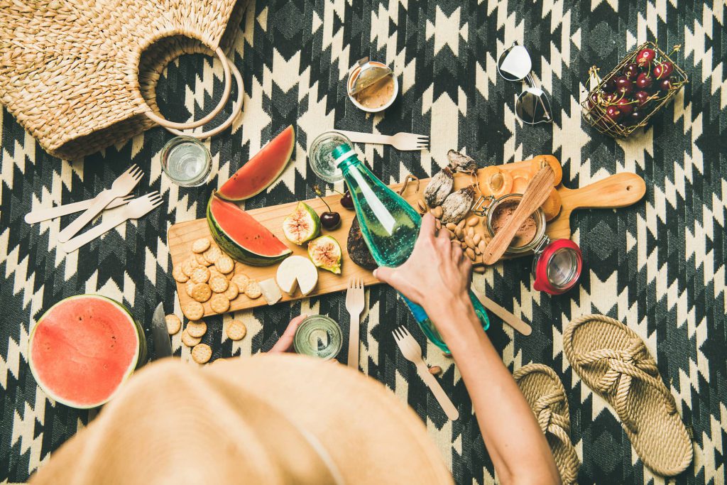 A woman pouring a sparkling water while feasting on a charcuterie board of nuts and fruits all on top of a hip southwestern design blanket.