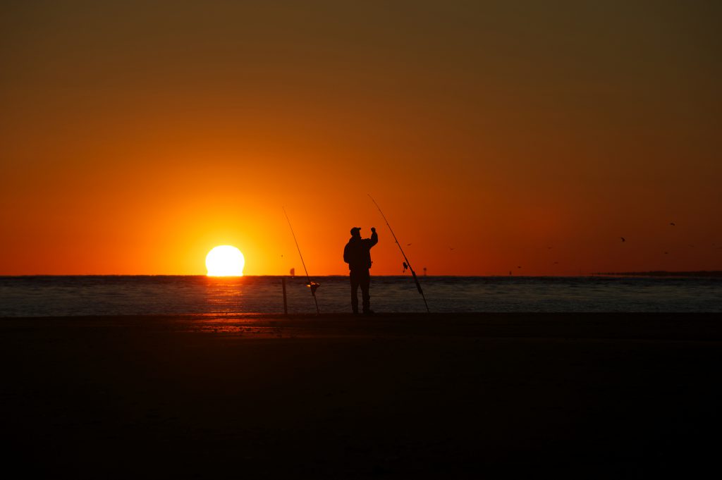 A fisherman is silhouetted by a sunset on a beach in North Carolina as he gets a rod ready to cast out. Birds can be seen flying in the background