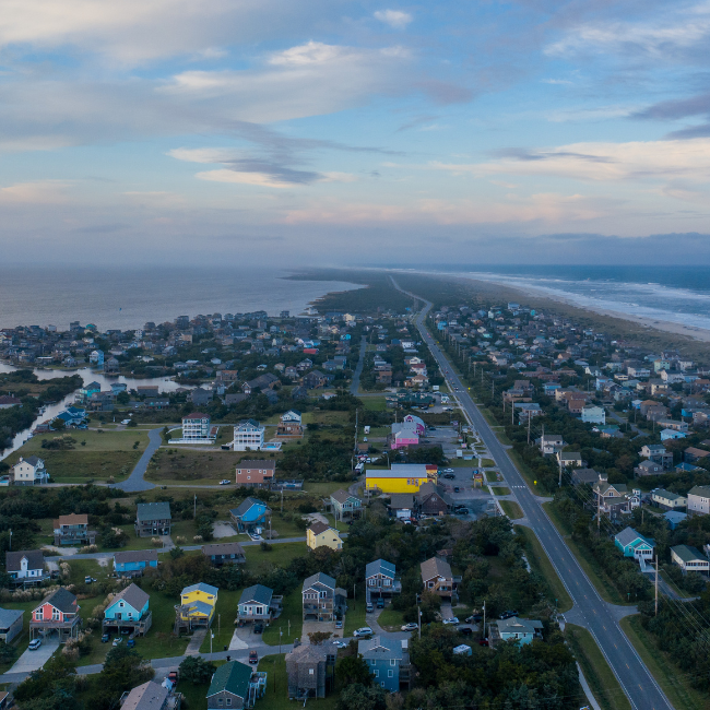 Aerial view of a coastal town at dusk with a main road running through the center, flanked by clusters of houses in various colors. 