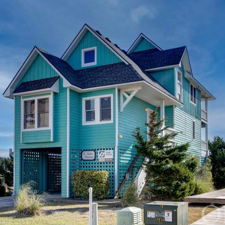 Frontal view of a two-story house painted in teal with white trims and a dark grey roof, featuring multiple windows and a staircase leading to an upper deck, situated next to a sandy pathway with shrubs on either side